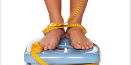 Woman’s feet on a scale with a measuring tape; she is calculating her weight loss.