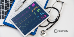 The image comprises of an ECG monitoring screen with ECG recordings superimposed against a backdrop of medical notes, laptop, stethoscope, and other equipment in a hospital/clinic facility that looks after population health.
