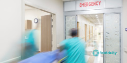 A team of healthcare specialists in scrubs wheel a stretcher into a room marked “Emergency.”